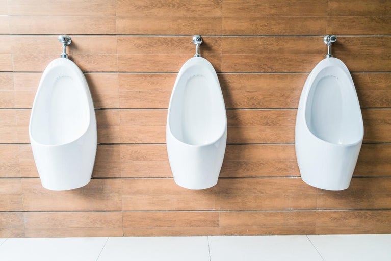 Trei pisoare suspendate din ceramică albă în fața unui perete cu plăci aspect lemn într-o baie modernă.