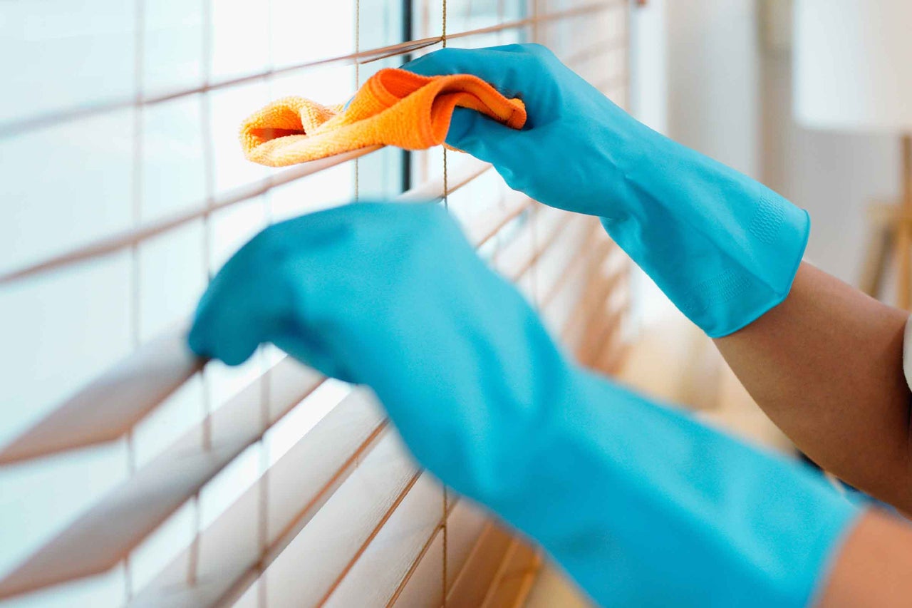 O persoană curăță jaluzelele din lemn cu o lavetă de curățare și mănuși de cauciuc.