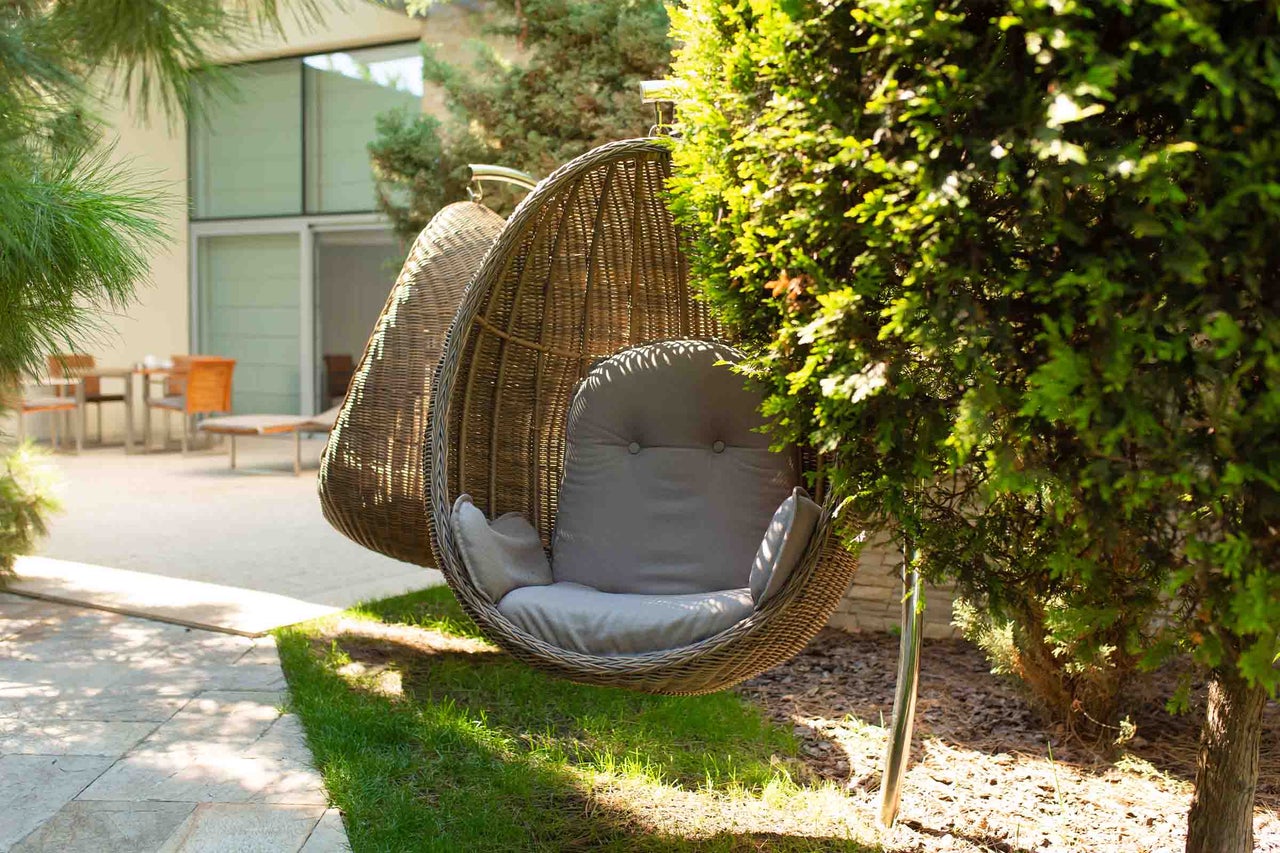Fotoliu suspendat din răchită în formă de ou cu perne într-o grădină luminoasă cu tufișuri și mobilier de terasă.