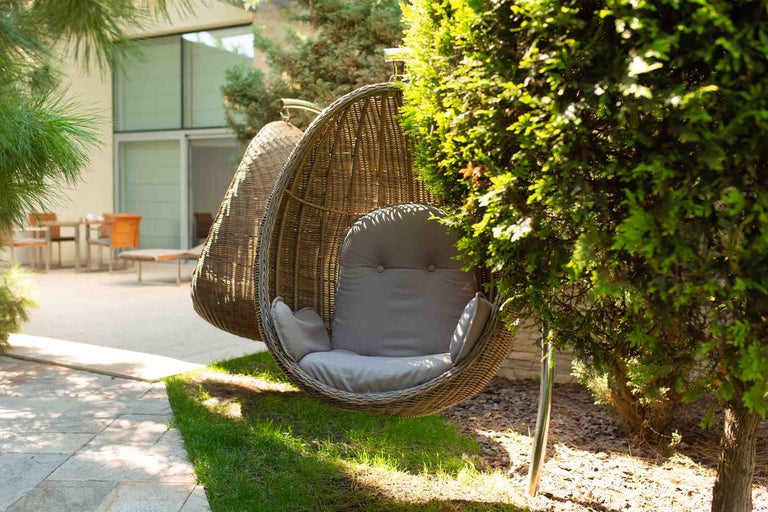 Fotoliu suspendat din răchită în formă de ou cu perne într-o grădină luminoasă cu tufișuri și mobilier de terasă.