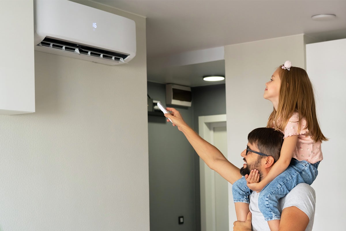 Un bărbat cu un copil pe umeri folosește o telecomandă pentru a regla un aparat de aer condiționat alb de perete într-un interior luminos.