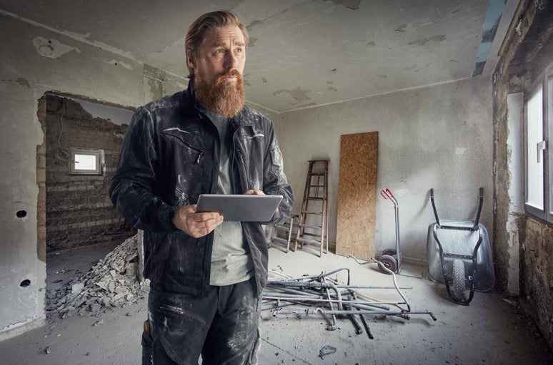 Meșter cu tabletă într-o cameră care are nevoie de renovare cu scară, roabă și țevi