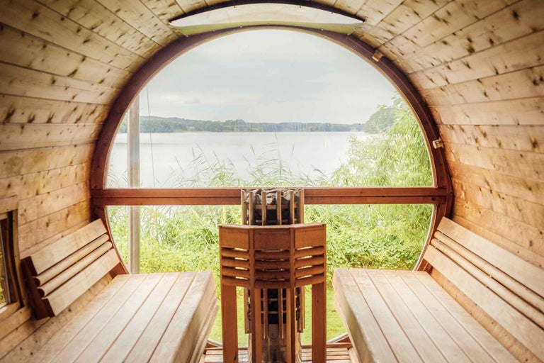 Vedere din interiorul unei saune de tip butoi din lemn cu fereastră rotundă mare spre un lac cu stuf și sobă de saună în prim plan.