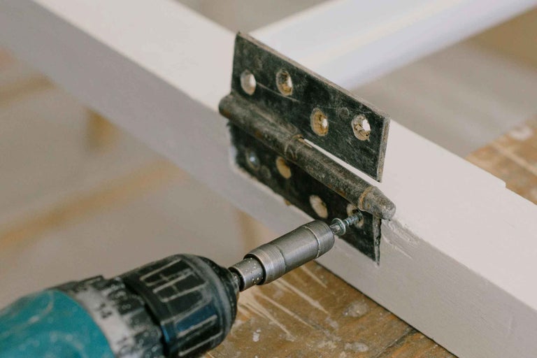 O șurubelniță cu acumulator fixează o balama pe un cadru de lemn.