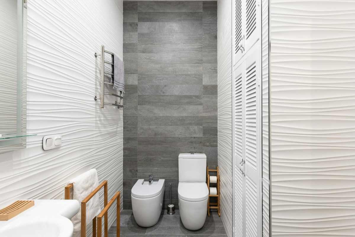 Baie modernă cu WC și bidet albe, gresie gri cu aspect de lemn și faianță albă cu structură ondulată.