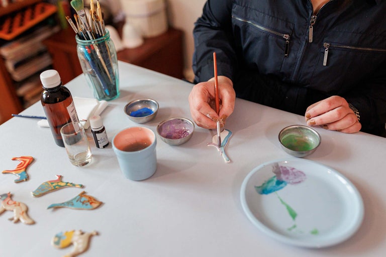 Scenă cu o persoană care pictează ornamente cu vopsele, pensule și diverse recipiente pe o masă, potrivite pentru activități artizanale.