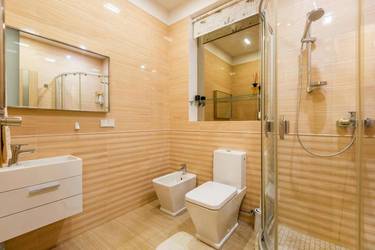 Baie luminoasă cu plăci bej, mobilier alb de lavoar, dulap cu oglindă, bidet, toaletă și cabină de duș din sticlă.