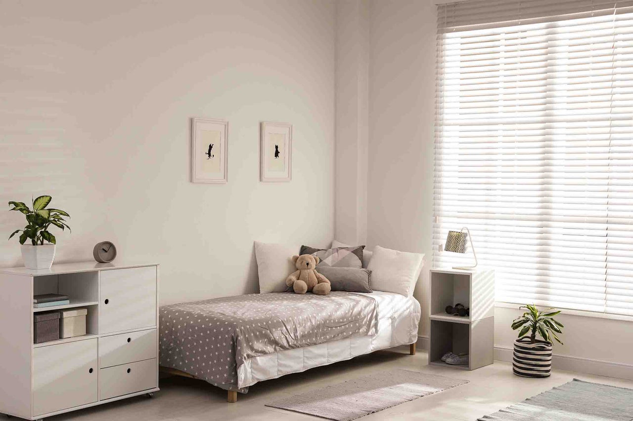 Cameră de copii luminoasă cu jaluzele orizontale albe la fereastră, un pat de o persoană cu imprimeu cu stele și o comodă albă.