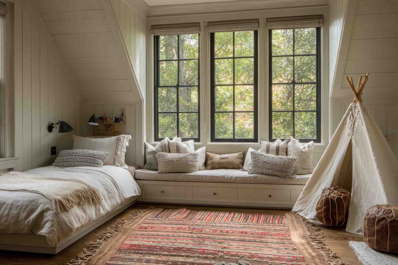 Cameră luminoasă la mansardă cu banchetă la fereastră, pat și cort tipi din pânză pe podea de lemn cu covor cu model.