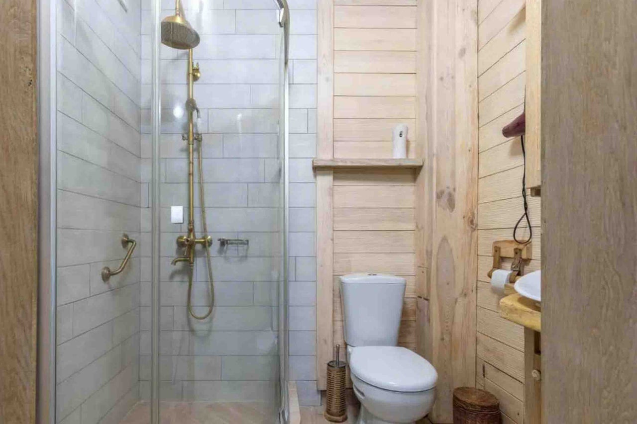 Baie compactă cu cabină de duș din sticlă, baterii din alamă, faianță deschisă la culoare, lambriu din lemn și vas WC alb pe pardoseală.