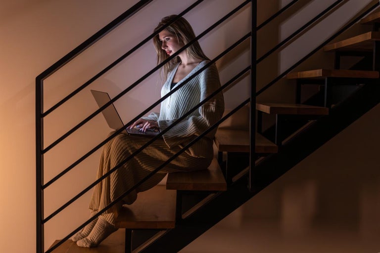 O femeie stă cu un laptop pe o scară cu balustradă metalică.