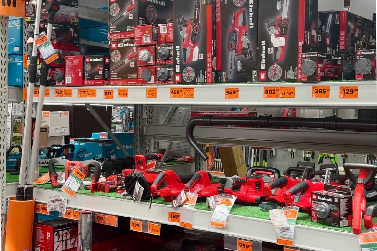 Raft într-un magazin de bricolaj cu diverse scule electrice marca Einhell, cum ar fi drujbe și suflante de frunze, cu etichete de preț.