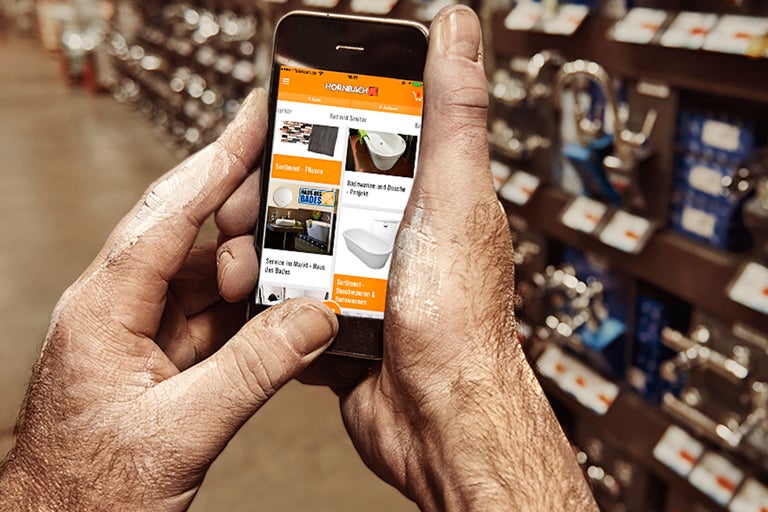 O mână ține un smartphone cu aplicația Hornbach deschisă în secțiunea de baie și sanitare