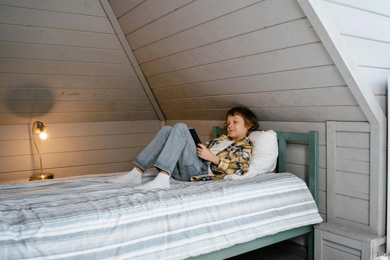 Băiat stând întins cu telefonul mobil pe un pat într-o cameră de mansardă.