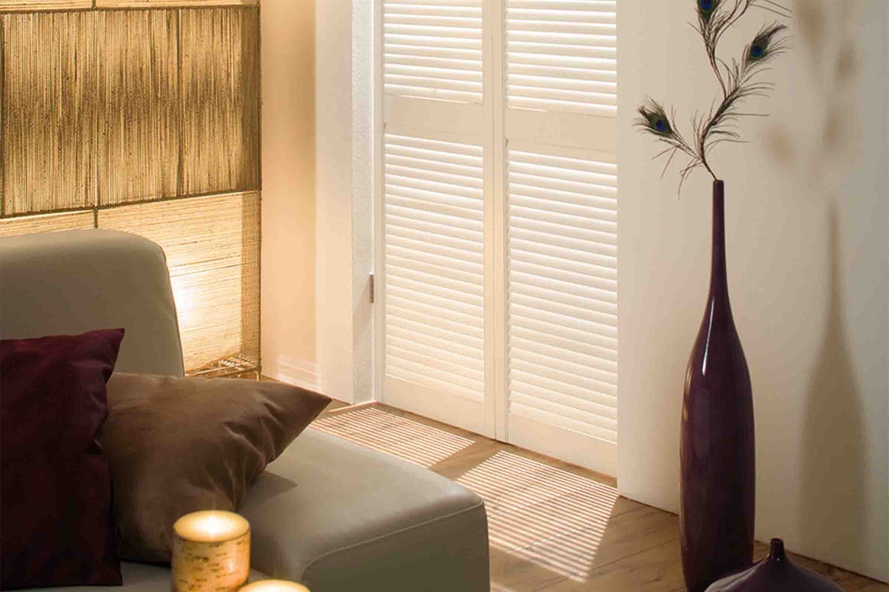 Uși albe cu lamele într-o cameră de zi luminoasă, cu o canapea, perne și o vază înaltă cu pene de păun.