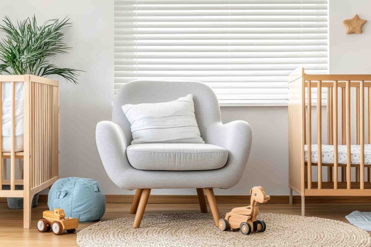 Cameră de copii luminoasă cu fotoliu gri pe picioare de lemn, două pătuțuri, jaluzele albe, covor rotund împletit și jucării din lemn.