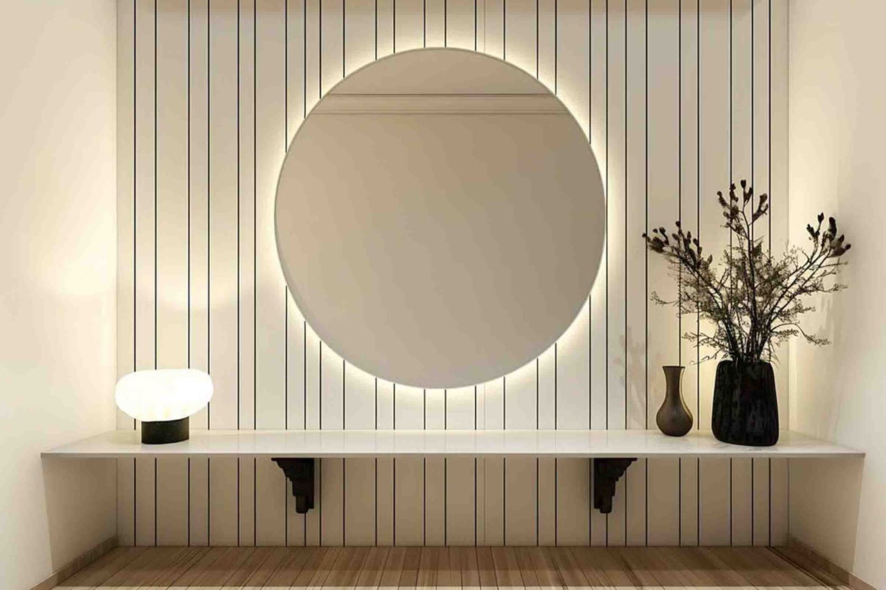 Cameră modernă cu oglindă rotundă iluminată din spate pe un perete în dungi, cu un raft alb dedesubt cu decorațiuni și lampă.
