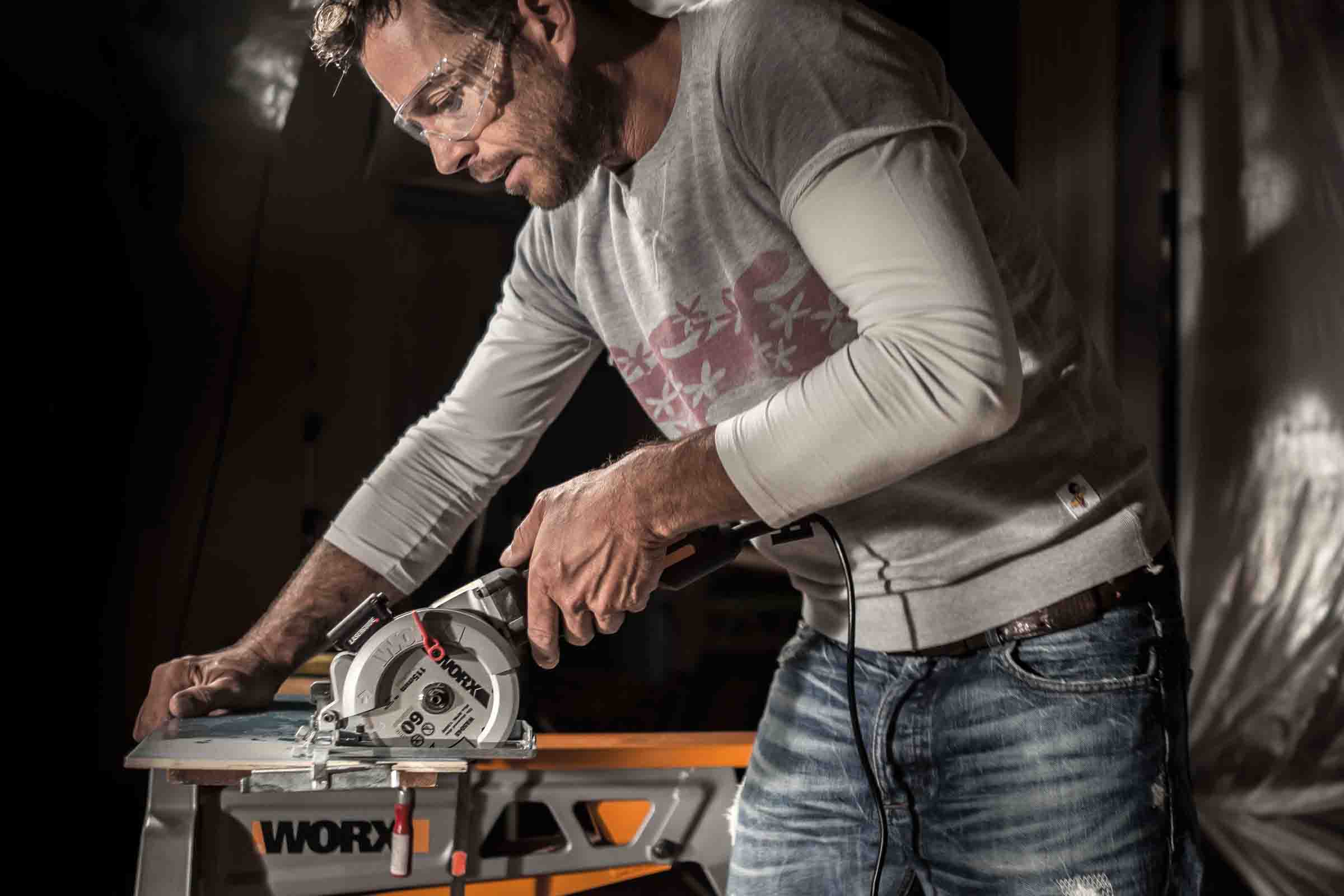 Un bărbat cu ochelari de protecție taie o piesă de lucru cu un ferăstrău circular portabil Worx pe un banc de lucru Worx.