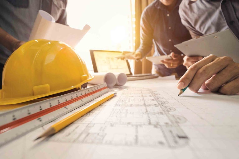 Cască de șantier, riglă, creion și plan de construcție pe o masă cu muncitori în construcții