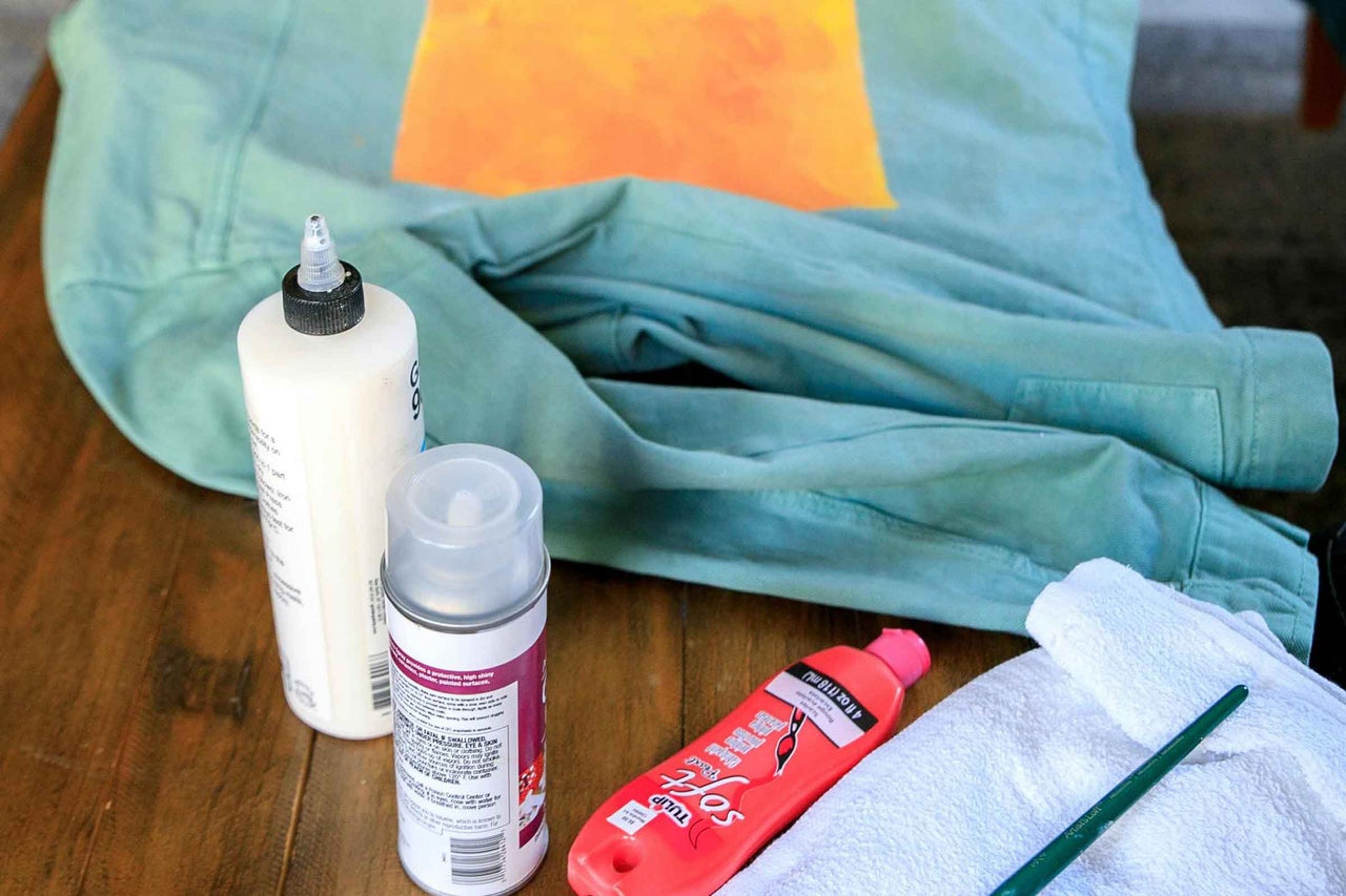 Fotografie cu un tricou cu vopsea textilă, adeziv, vopsea spray, pensulă și prosop.
