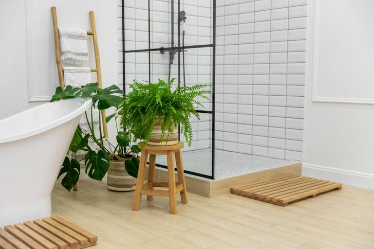 Baie modernă luminoasă cu cadă albă, duș de tip walk-in cu ramă neagră, accesorii din lemn și plante de interior verzi.