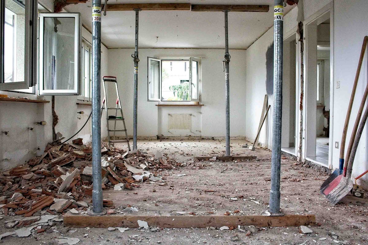 Cameră în renovare cu popi metalici care susțin o grindă de lemn, moloz pe podea și o scară dublă.