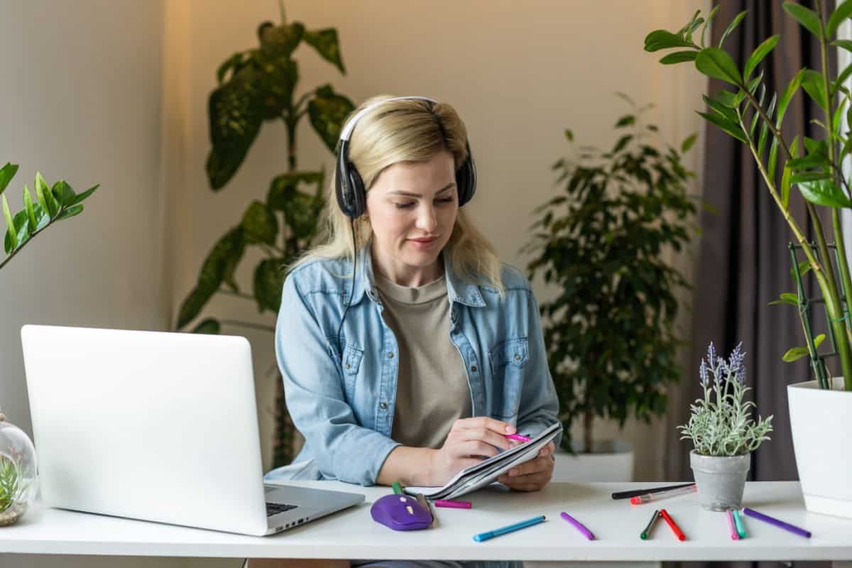 Femeie cu căști care își ia notițe la un birou cu un laptop și plante.
