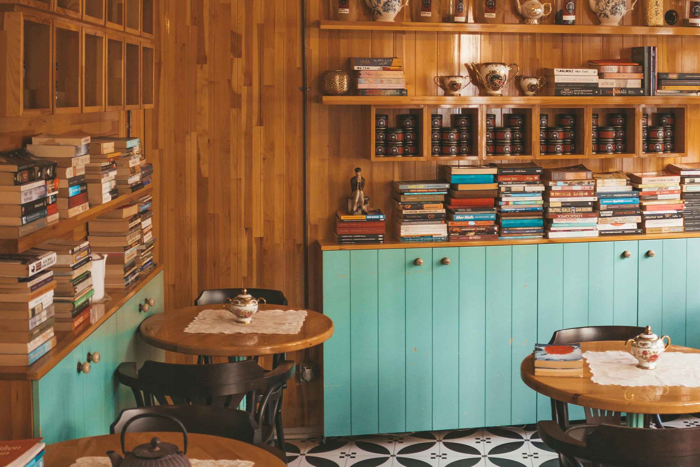 Vedere interioară a unei cafenele sau biblioteci cu pereți din lemn, rafturi cu cărți și ceainic, precum și mese cu scaune.