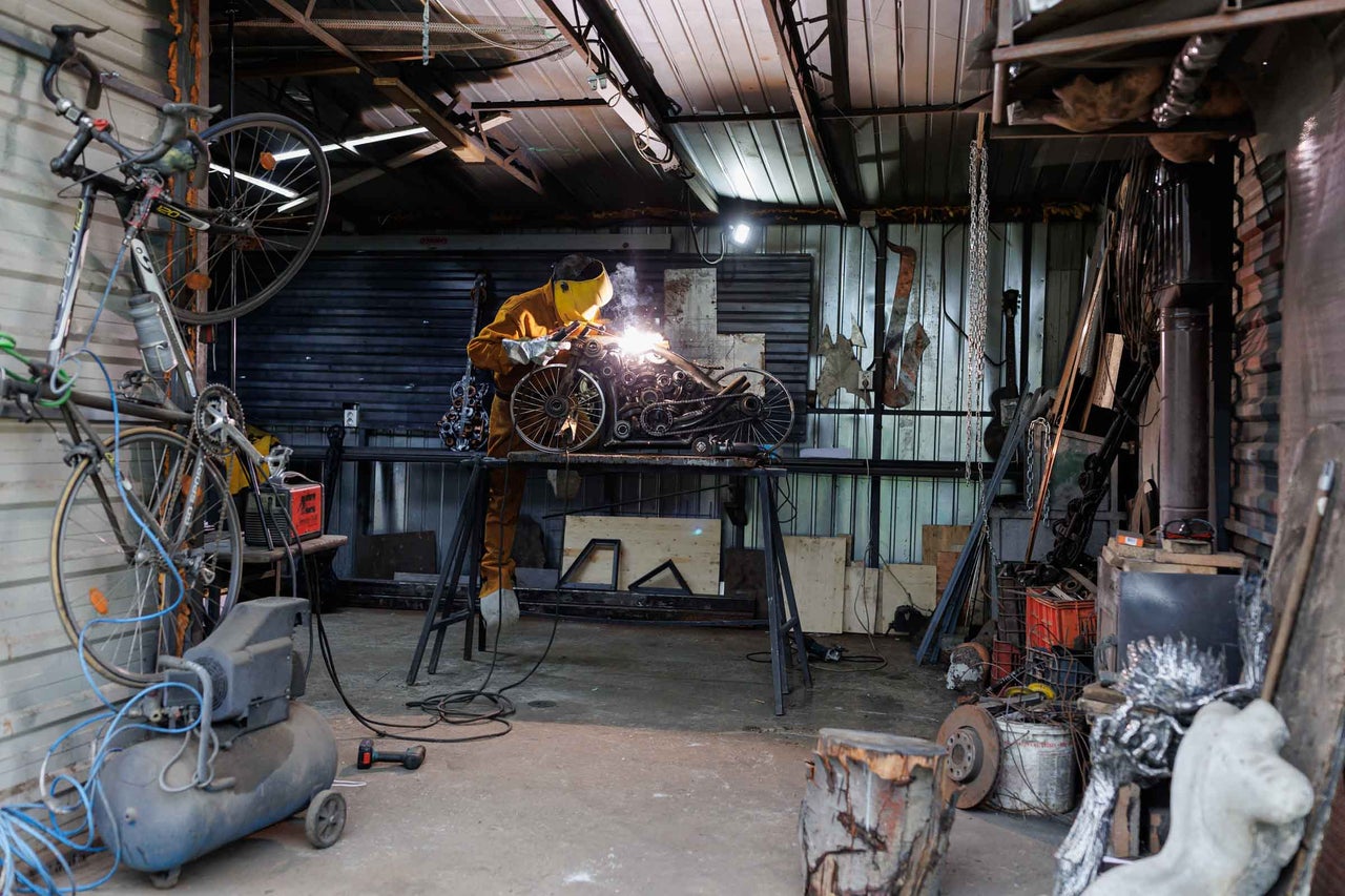 Un artizan sudează o sculptură metalică într-un atelier, înconjurat de biciclete, scule și echipamente.
