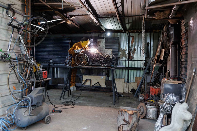 Un artizan sudează o sculptură metalică într-un atelier, înconjurat de biciclete, scule și echipamente.
