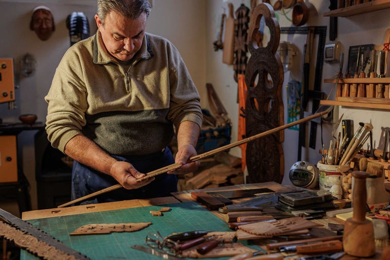 Un bărbat lucrează la un ornament decorativ din lemn în atelierul său. Pe masă sunt așezate unelte și sculpturi în lemn.