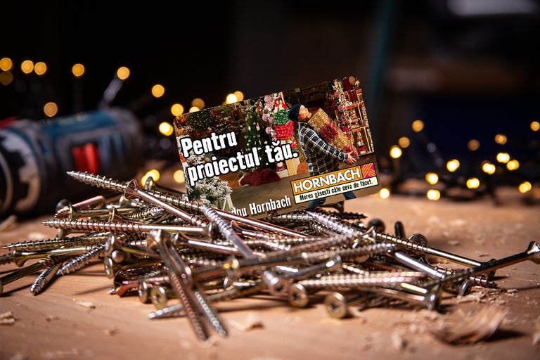 Prim-plan cu șuruburi cu card cadou Hornbach și mașină de găurit în fundal.