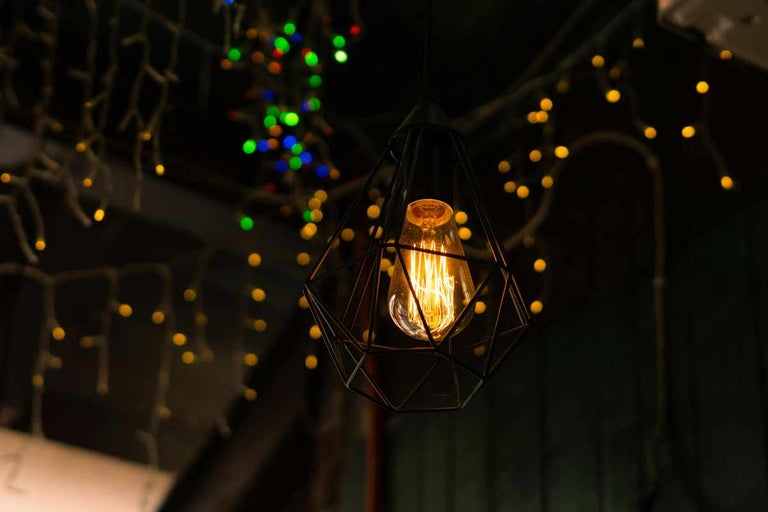 Lustră din metal negru cu abajur tip cușcă geometrică și bec decorativ cu filament.
