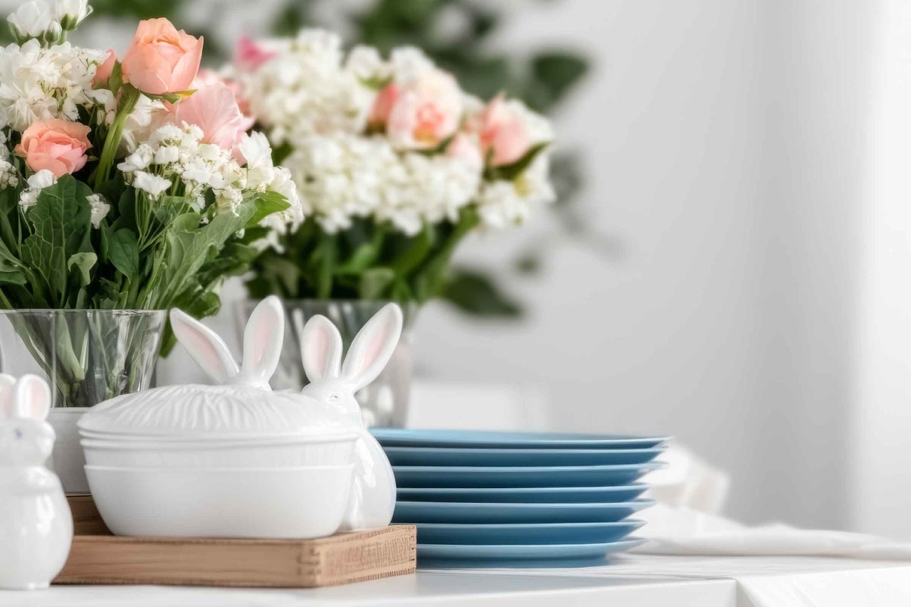 Decorațiune luminoasă de Paște cu cutii din porțelan în formă de iepure, o stivă de farfurii albastre și buchete de flori în vaze.