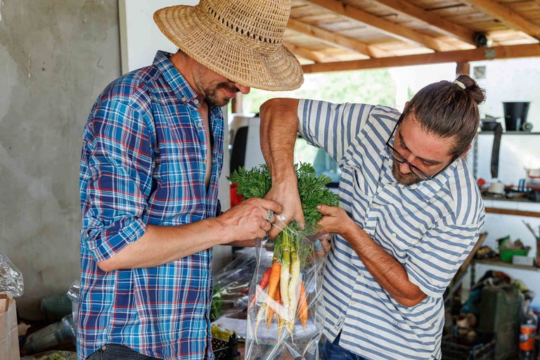 Doi bărbați ambalează un mănunchi de morcovi într-o pungă de plastic.