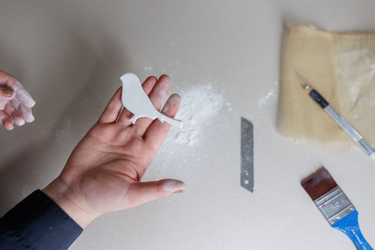 O mână ține o formă albă de pasăre lângă unelte de artizanat