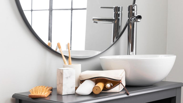 Lavoar modern cu chiuvetă pe blat albă, baterie cromată, oglindă rotundă și accesorii de baie din lemn și ceramică.