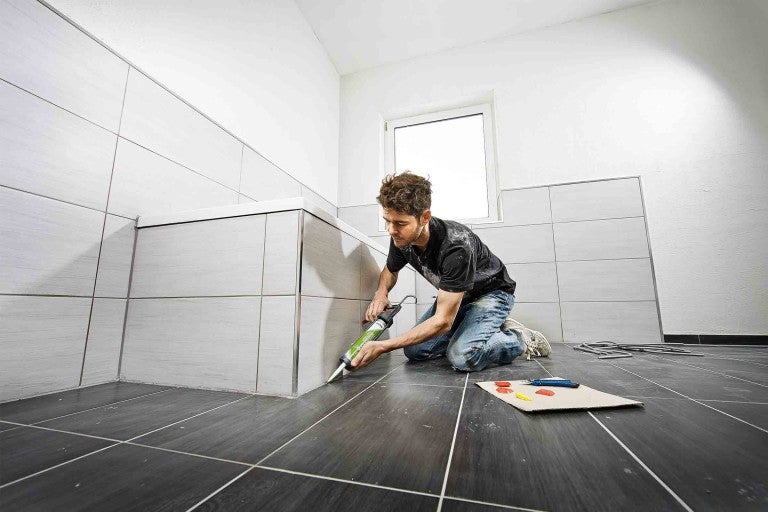 Meșterul chituiește plăcile cu un pistol cartuș în baie.