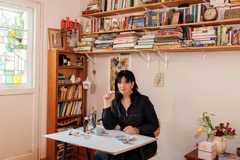 Un birou luminos cu rafturi de cărți și o femeie așezată la o masă pictând.