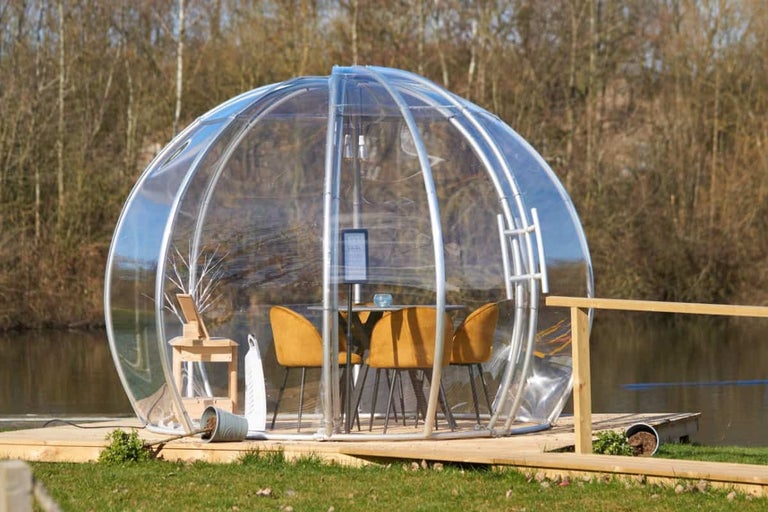 Pavilion exterior transparent cu masă și scaune pe o terasă de lemn lângă apă