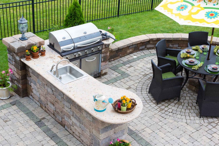 Bucătărie de exterior cu grătar, chiuvetă și masă cu scaune pe o terasă în grădină.