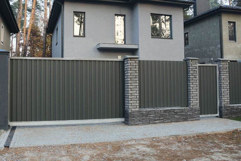 Împrejmuire modernă a terenului cu o poartă glisantă mare din metal profilat și o poartă de grădină potrivită între stâlpi de piatră.