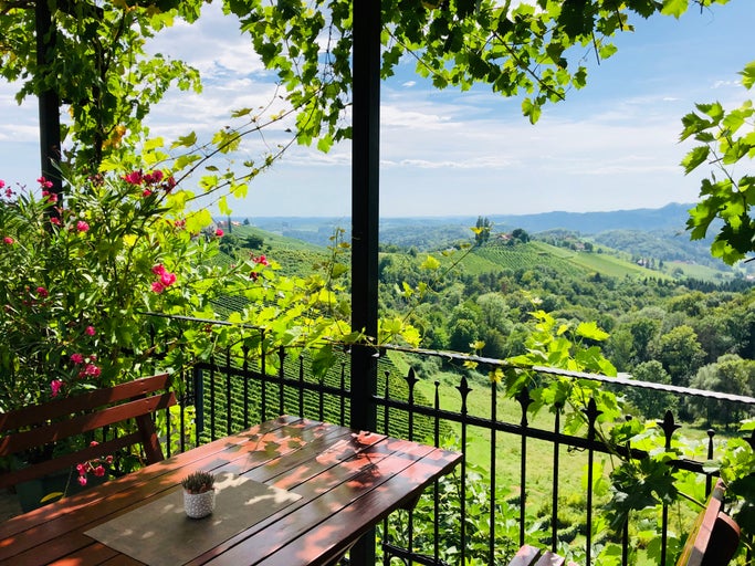 Vedere de pe o terasă cu masă de lemn și balustradă metalică spre un peisaj deluros