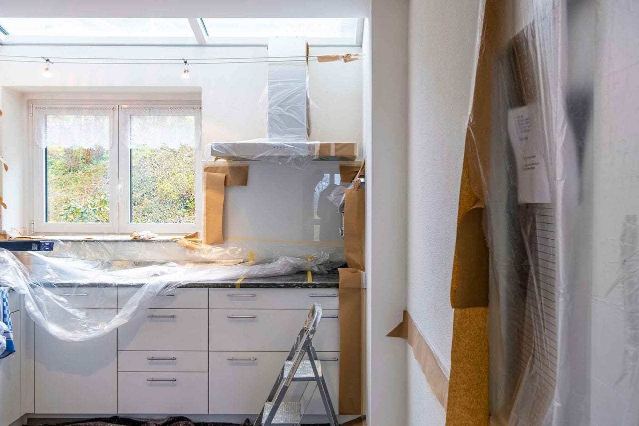 Bucătărie albă în curs de renovare, dulapurile și blatul fiind acoperite cu folie de protecție și hârtie de mascare, cu o scară în față.