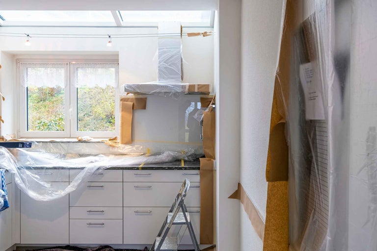 Bucătărie albă în curs de renovare, dulapurile și blatul fiind acoperite cu folie de protecție și hârtie de mascare, cu o scară în față.