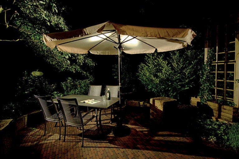 Masa de grădină iluminată cu umbrelă de soare, scaune și plante în grădină pe timp de noapte.