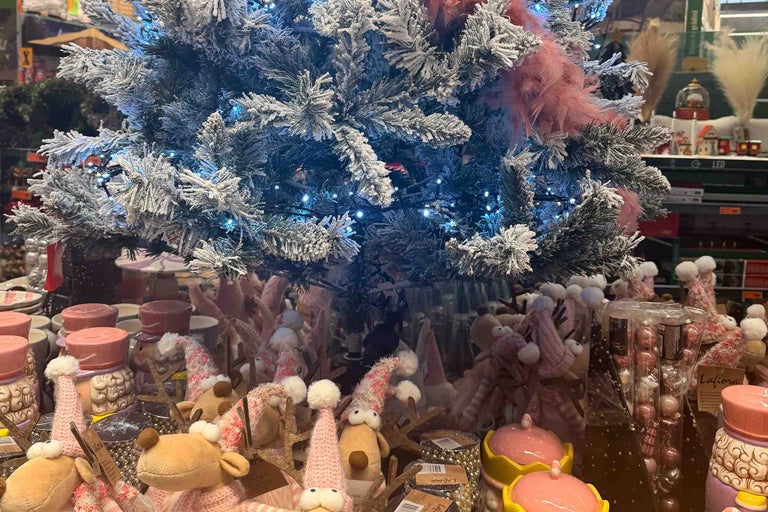 Decor de Crăciun în magazin cu brad artificial înzăpezit, ghirlandă luminoasă LED și diverși reni de pluș și accesorii decorative.
