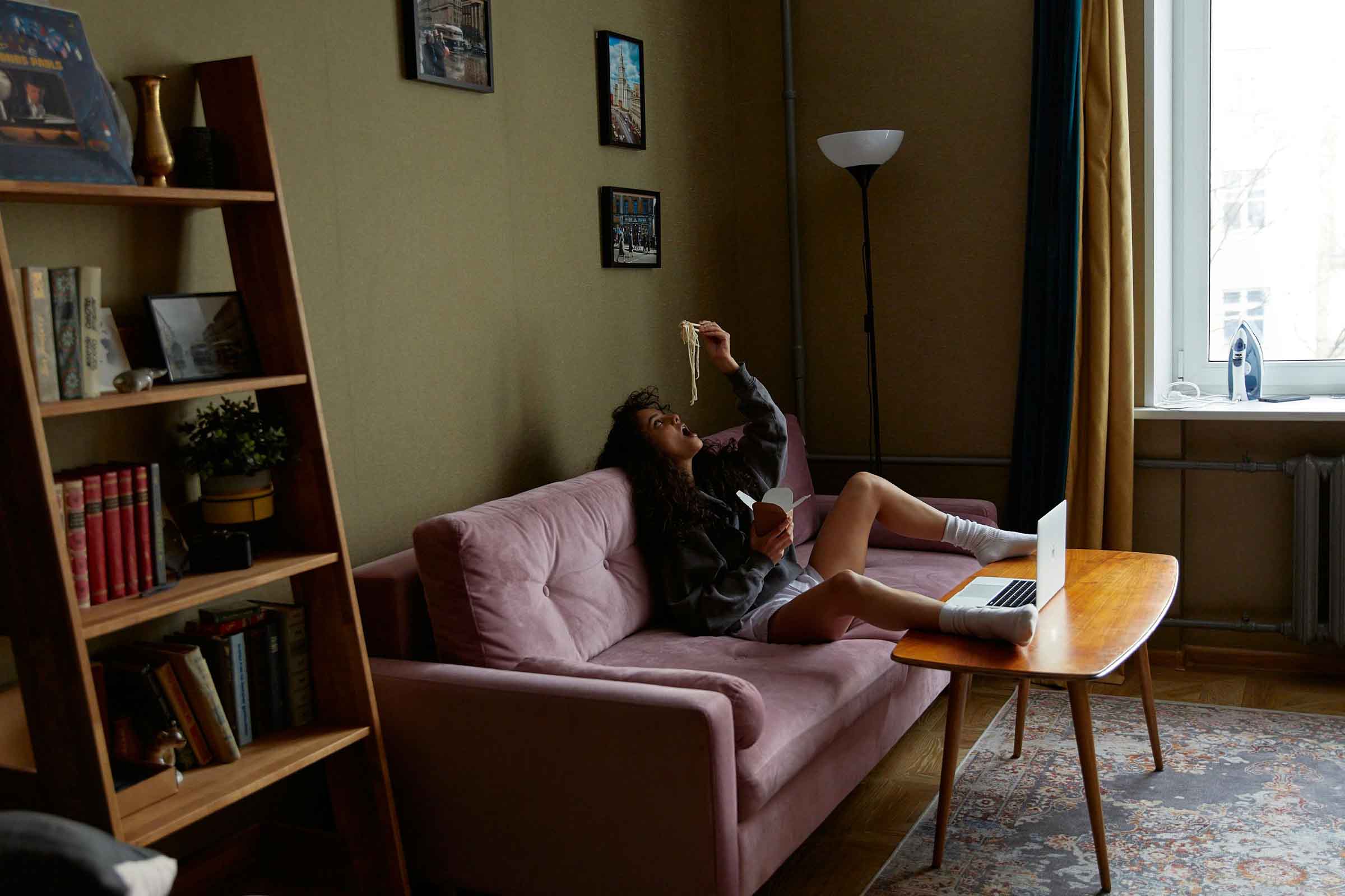 Scenă de sufragerie cu o femeie întinsă pe o canapea în timp ce mănâncă tăiței și folosește un laptop, cu o bibliotecă și o masă în cameră