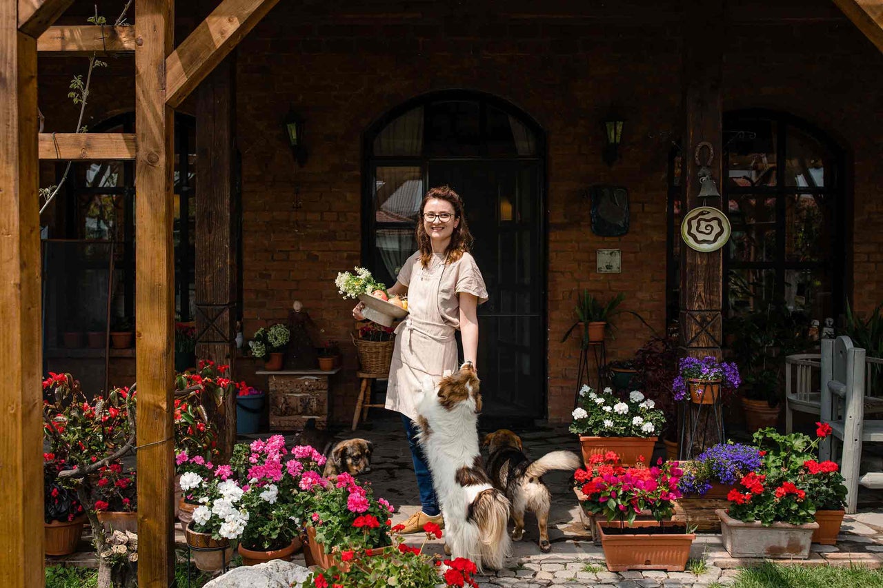 Femeie cu flori în fața unei case cu jardiniere cu flori și câini.