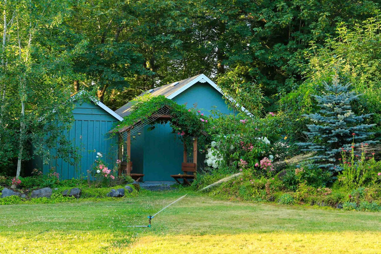 Două căsuțe de grădină din lemn albastru într-un peisaj verde cu flori, gazon și un aspersor activ.